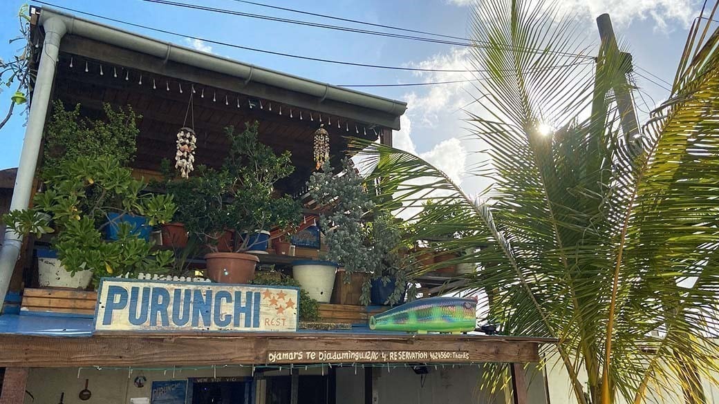 The balcony of the Purunchi Fisherman's House, planted with large potted plants - with wind chimes, name plate, opening hours and telephone number