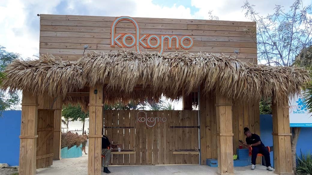 Einladender Eingang des Kokomo Beach, der wie eine mit Palmen bedeckte Holzhütte aussieht, mit großem Logo und zwei der Schaukeln, für den der Strand bekannt ist